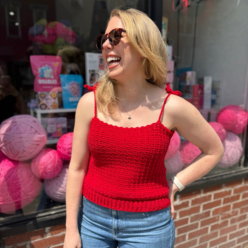 A blonde woman wearing sunglasses and a red crochet tank top with bow straps.