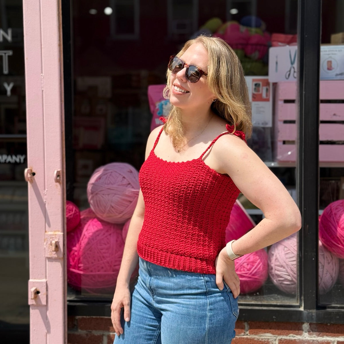 A blonde woman wearing sunglasses and a red crochet tank top with bow straps.