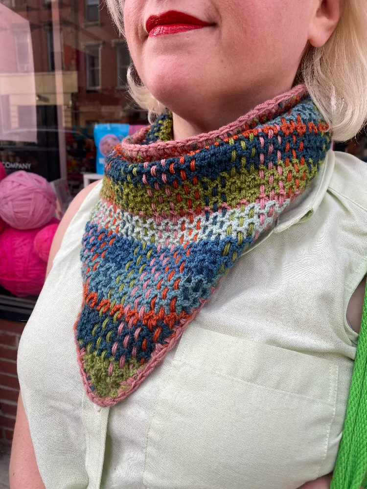 A close up shot of a woman with red lipstick wearing a plaid crochet kerchief tied around her neck.